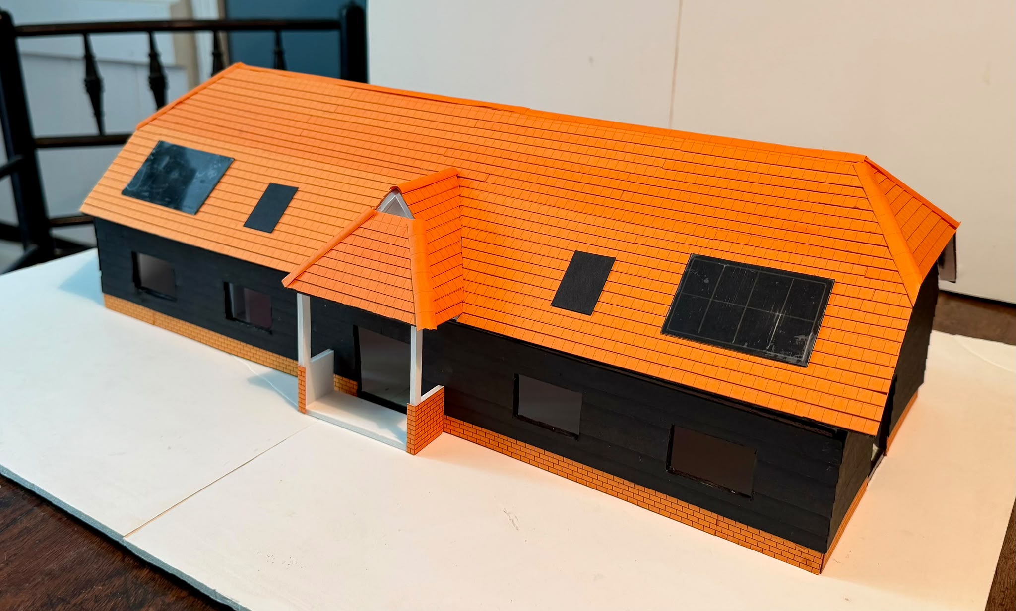 A card model of the new village hall: a long building with black timber-style walls, an orange tiled roof with solar panels and roof windows, and a small porch entrance at the front