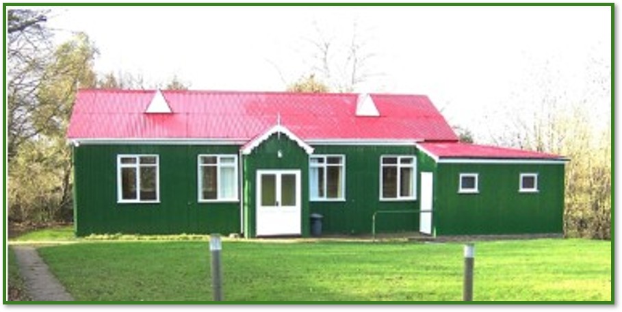 Hever Village Hall, a green timber-clad hall with a red roof, seen from the front lawn