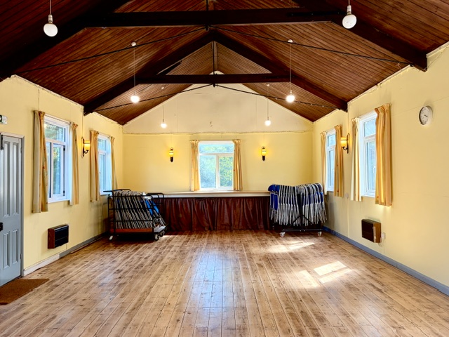 Inside the main hall at Hever Village Hall: a wood-floored room with a high pitched ceiling, stacked chairs and a stage at the far end